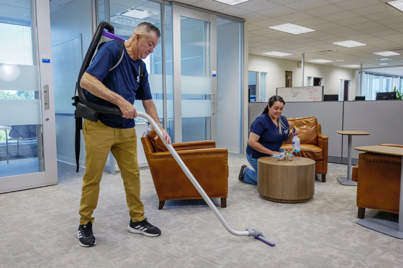 An ExtraClean team member vacuuming an office lounge while a coworker wipes a table